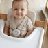 Baby in a high chair with a beige harness, smiling.